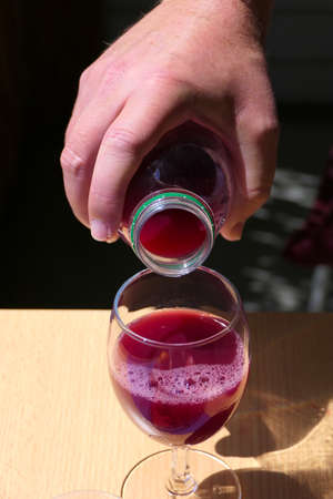 Closeup hand pouring out red fruit juice or pomegranate juice in a bottle into a glass on a wooden tableの写真素材