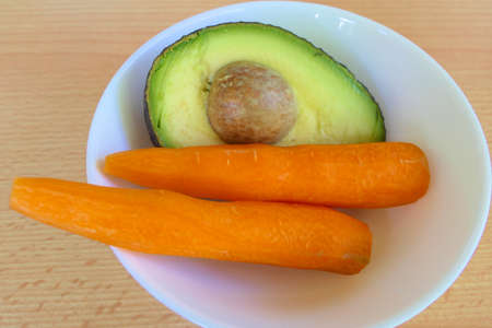 Healthy juice ingredients - closeup half avocado and carrots in a white bowl on a tableの写真素材