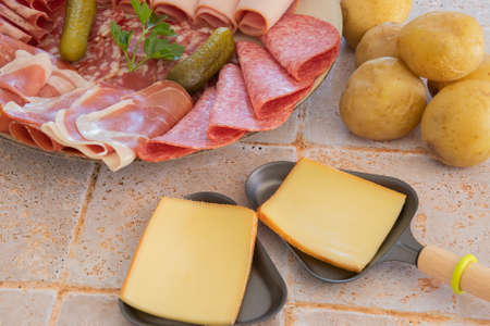 raw raclette cheese in pans and cold cuts on a table with potatoesの写真素材