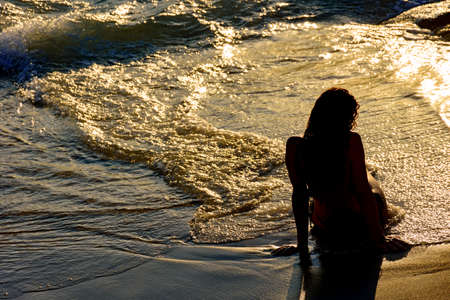 Girl sitting on the sand near the water during sunset in summer
