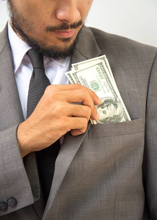 Wealthy handsome. Close-up of young man in putting money in his pocket while standing against grey backgroundの写真素材