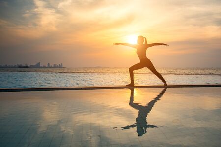 Silhouette young woman practicing yoga on swimming pool and the beach at sunsetの写真素材