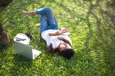 Girl enjoy listening music and reading a book and play laptop on the grass field of the park in the morning (greenery tone)の写真素材