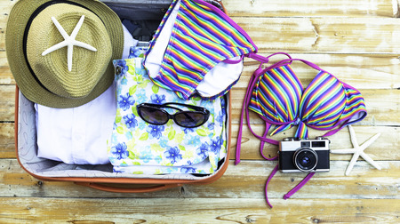 Open the suitcase with tourist things: women's hat, swimsuit, camera, denim shorts, dresses, sunglasses, perfumes, nail polish, mobile phone, tablet on wooden backgroundの写真素材