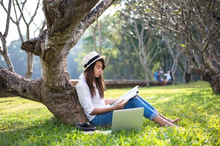 Girl enjoy reading a book under the tree, laying on grass of park outdoor in morningの写真素材