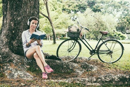 Woman reading a book while listening music in a parkの写真素材