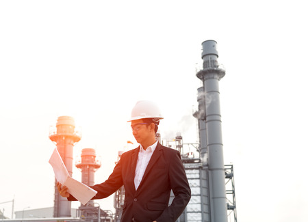 Engineer wearing white helmet holding paper drawing with oil refinery background.の写真素材