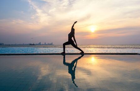Silhouette young woman practicing yoga on swimming pool and the beach at sunset.の写真素材