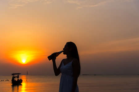 Silhouette woman raised hands holding beer bottle on the sunset skyの写真素材