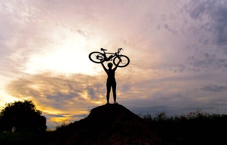 Silhouette the women in action lifting bicycle above his head stand on meadow with sunsetの写真素材