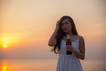Silhouette woman raised hands holding beer bottle on the sunset skyの写真素材