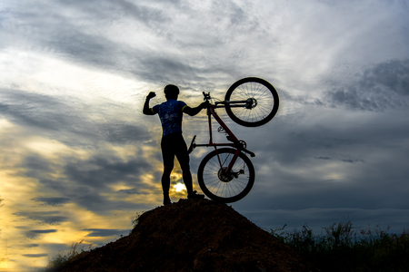 Silhouette the man in action lifting bicycle above his head stand on meadow with sunsetの写真素材