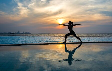 Silhouette young woman practicing yoga on swimming pool and the beach at sunset.  Healthy Concept.の写真素材