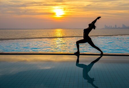Silhouette young woman practicing yoga on swimming pool and the beach at sunset.  Healthy Concept.の写真素材