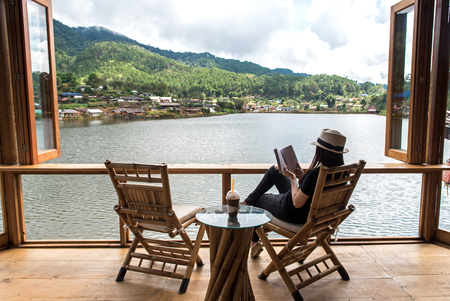 Woman reading a book lying and drink ice coffee on the chair in wood terrace aginst beautiful veiw, ralax and happy day.  Lifestyle Conceptの写真素材
