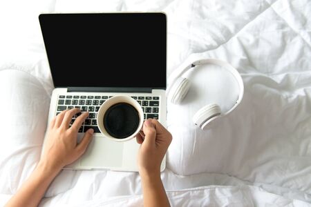Male sitting on white bed surfing internet or working on laptop and listening to musical media online with wireless headphone and drink coffee in morning relax mood. Music and Lifestyle Concept.の写真素材