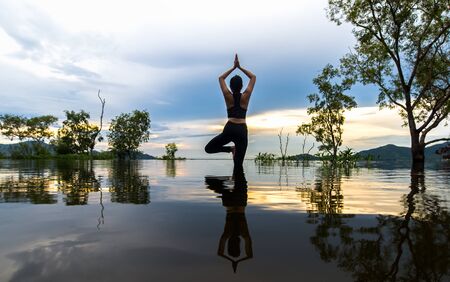 Silhouette young woman lifestyle exercising vital meditate and practicing reflect on Flood the trees in the reservoir, background sunset.  Healthy Concept.の写真素材