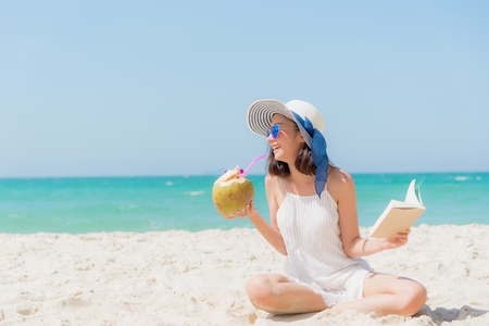 Summer Vacation. Smelling asian women relaxing, reading book and drinking coconut cocktail on the beach, so happy and luxury in holiday summer, blue sky background.  Travel and lifestyle Concept.の写真素材