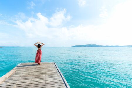 Summer Day. Smiling women relax and wearing red dress fashion standing on the wooden bridge over the sea, blue sky background. Travel and Vacation. Freedom Conceptの写真素材