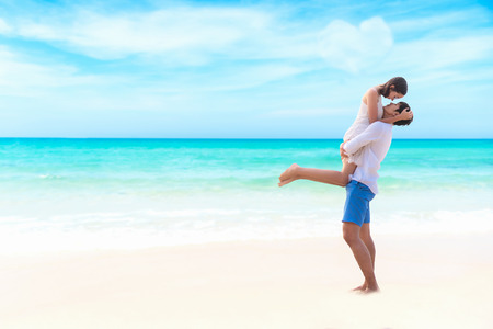 Couple in love. Smiling asian young man is holding girlfriend in his arms on the beach on evening time, blue sky heart shape background.   Honeymoon relax together on summer travel.  Valentine, Travel and Summer Concept.の写真素材