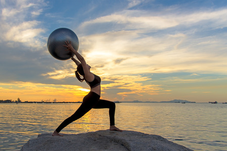 Silhouette  healthy woman lifestyle exercising vital meditate and practicing yoga and gym ball on the rock at beach sunset and twilight time, city night background.  Healthy Concept.の写真素材