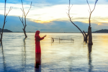Veiled Islamic muslim woman wearing a burka standing and praying in the river, sunset backgroundの写真素材