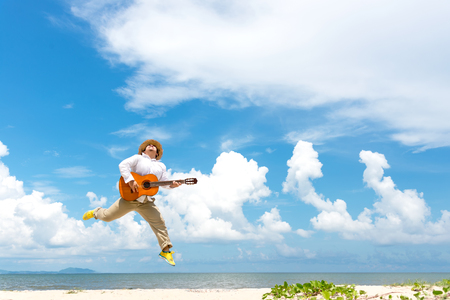 Asian man enjoying summer, playing classic guitar and jumping on the beach in vacations, blue sky background.  Travel and Summer Conceptの写真素材