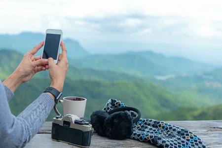 Traveler woman using smartphone and drinking  coffee and tea and take a photo and relax in sun sitting outdoor in sunshine light enjoying her warm morning at balcony house, winter season. Lifestyle Conceptの写真素材