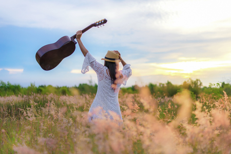 Lifestyle women white dress holding a guitar on a cloudy sunset or sunrise sky in the meadow flower, relax and happy day on summertime,  background field copy space for inscription.  Lifestyle and Vacation Conceptの写真素材