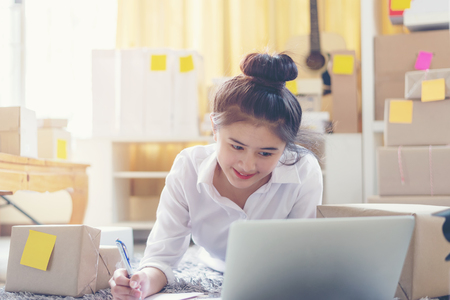 Asian smiling young women writing order from customer online for preparing package product.  Lifestyle for working at home.の写真素材