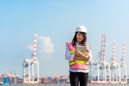 Asian women engineer holding laptop and working with container Cargo freight ship in shipyard at dusk for Logistic Import Export background, safety control. Engineer Conceptの写真素材