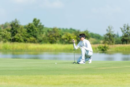 Professional Golfer asian man playing aiming shot for putting ball on the hole with club on green course.  Hobby in holiday and vacations in sunny morning day on multiethnic club golf. Lifestyle and Sport Conceptの写真素材