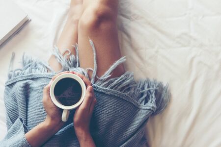 Close up legs women on white bed in the bedroom. Women reading book and drinking coffee in morning relax mood in winter season. Lifestyle Concept.の写真素材