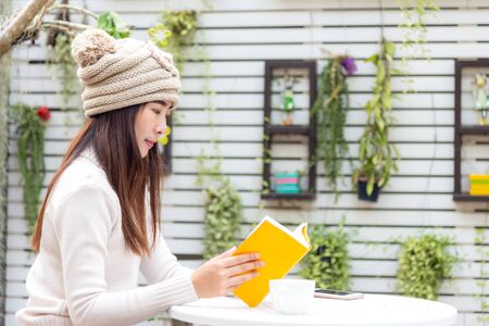 Asian young woman wearing sweater, sitting and reading book with hot coffee, smart phone at home garden in winter season. Lifestyle Conceptの写真素材