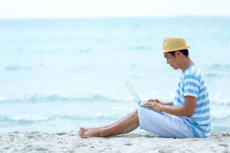 Lifestyle young asian man working on laptop while sitting chill on the beautiful outdoor beach, freelance working social on holiday summer. Summer and Vacation Conceptの写真素材