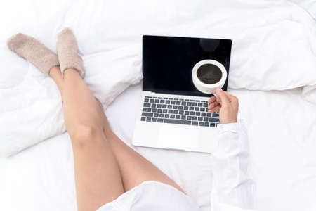 Close up legs women on white bed in the bedroom. Women working on laptop and drinking coffee in morning relax mood in winter season. Lifestyle Concept.の写真素材