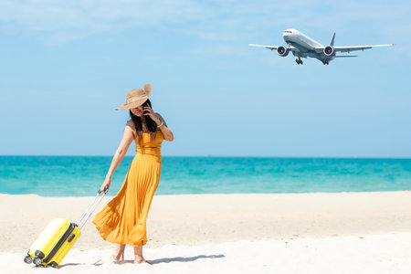 Asia women traveler holding yellow luggage walking and check in smart phone on the beach. Traveling on an airplane for leisure and destination.  Traveler and Tourism planning trips summer vacations. Summer Conceptの写真素材