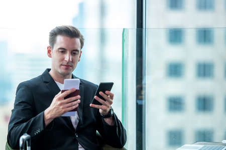 Businessman holding passport and  using smartphone check in air ticket  and boarding card in Departure Lounge at terminal airport.  People tourism business sitting working tablet computer and wait the airplane at the airport glass window for destination leisure.の写真素材