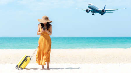 Asia women traveler holding yellow luggage walking and check in smart phone on the beach.  Traveling on an airplane for leisure and destination.  Traveler and Tourism planning trips summer vacations. Summer Conceptの写真素材