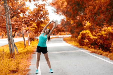Healthy woman warming up before jogging run and relax stretching her arms and looking away in the road outdoor. Asian runner people workout fitness session, autumn nature park background. Healthy and Lifestyle Concept.の写真素材