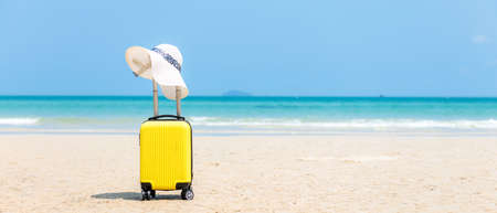 Summer traveling and tourism planning with yellow suitcase luggage with big hat fashion in the sand beach. Travel in the holiday, blue sky and beach background. Summer and Travel Concept, copy space for bannerの写真素材