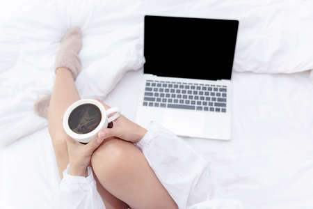 Close up legs women on white bed in the bedroom. Women working on laptop and drinking coffee in morning relax mood in winter season. Lifestyle Concept.の写真素材