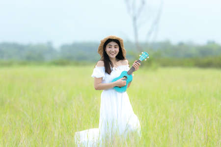 Lifestyle asian women white dress holding a guitar on a cloudy sunrise sky in the meadow flower, relax and happy day on summertime, outdoors background field copy space for inscription.の写真素材