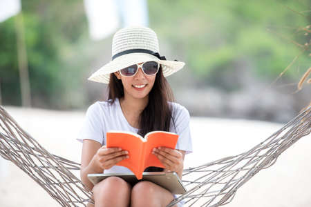 Lifestyle young woman using laptop working and reading book for relax on the beach.Â  Asian smiling people on hammock success and together your work pastime and meeting conference on internet in holidayの写真素材