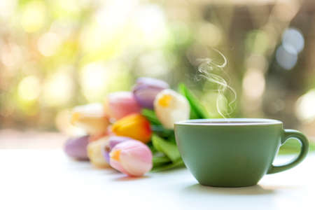 Green steaming cup of hot coffee on white wooden near window with tulip flowers and green nature blurred background.の写真素材