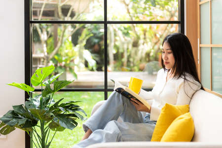 soft focus. Happy young woman reading book on sofa at home.  Lifestyle freelance relax and chill drinking coffee in living room, Nature garden background.  Lifestyle Conceptの写真素材