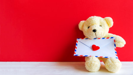 Cute teddy bear clutching love letter in its arms on wooden red background, copy space. Valentine Conceptの写真素材