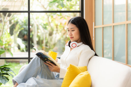 Happy young woman reading book on sofa at home.  Lifestyle freelance relax and chill drinking coffee in living room, Nature garden background.  Lifestyle Conceptの写真素材