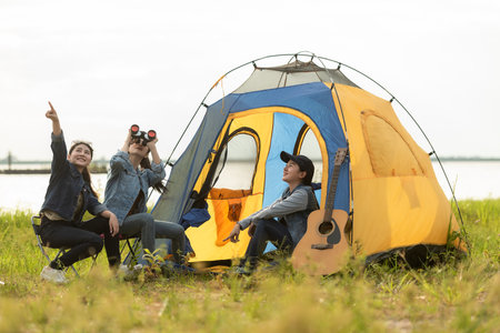 Camping outdoor. Group  friends camping leisure and destination travel. Family sitting around camp fire and playing guitar and roasted sausages, tourism relax and travel near river in holiday.の写真素材