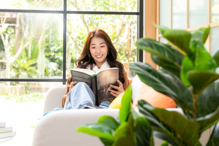 Happy young woman reading book on sofa at home.  Lifestyle freelance relax and chill drinking coffee in living room, Nature garden background.  Lifestyle Conceptの写真素材